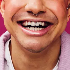 Uśmiechnięta twarz z kolczykiem w nosie i zębami ozdobionymi biżuterią. Widoczny frenum piercing dodaje charakteru.