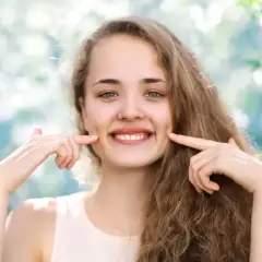 Uśmiechnięta dziewczyna z dołeczkami na policzkach, które podkreśla palcami. Wygląda jakby miała cheek piercing, ale to naturalne wgłębienia.