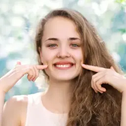 Uśmiechnięta dziewczyna z dołeczkami na policzkach, które podkreśla palcami. Wygląda jakby miała cheek piercing, ale to naturalne wgłębienia.