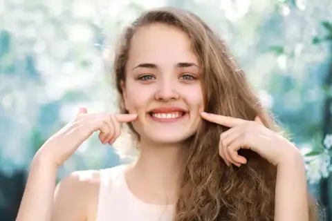 Uśmiechnięta dziewczyna z dołeczkami na policzkach, które podkreśla palcami. Wygląda jakby miała cheek piercing, ale to naturalne wgłębienia.