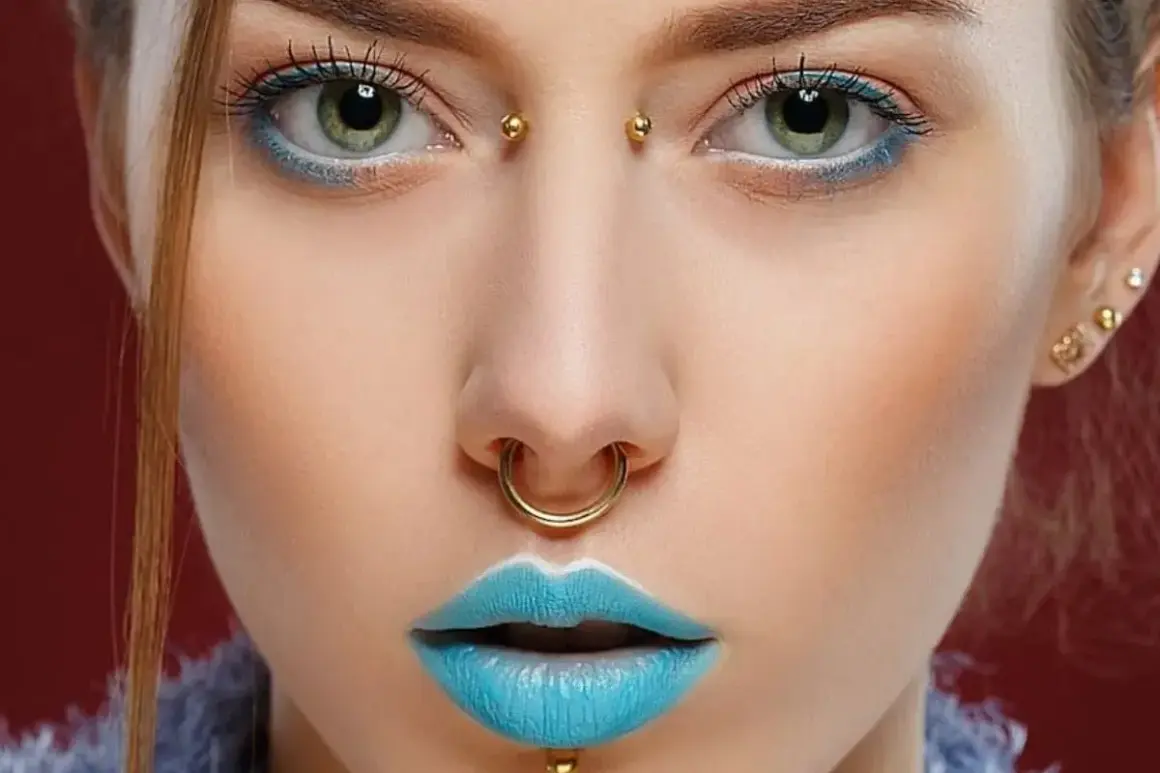 Dziewczyna z zielonymi oczami, błękitnymi ustami i złotymi kolczykami: septum, bridge piercing i w uchu.