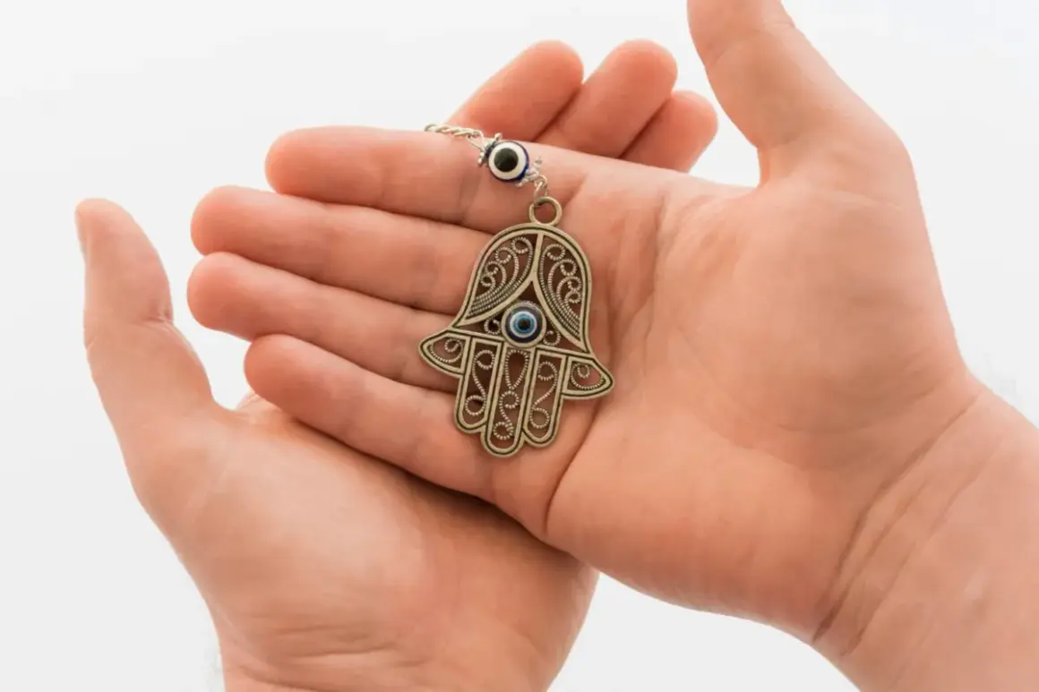 Dłonie trzymają ozdobny amulet ręka Fatimy z niebieskim okiem, symbol ochrony.