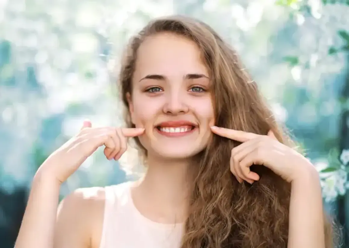 Uśmiechnięta dziewczyna z dołeczkami na policzkach, które podkreśla palcami. Wygląda jakby miała cheek piercing, ale to naturalne wgłębienia.