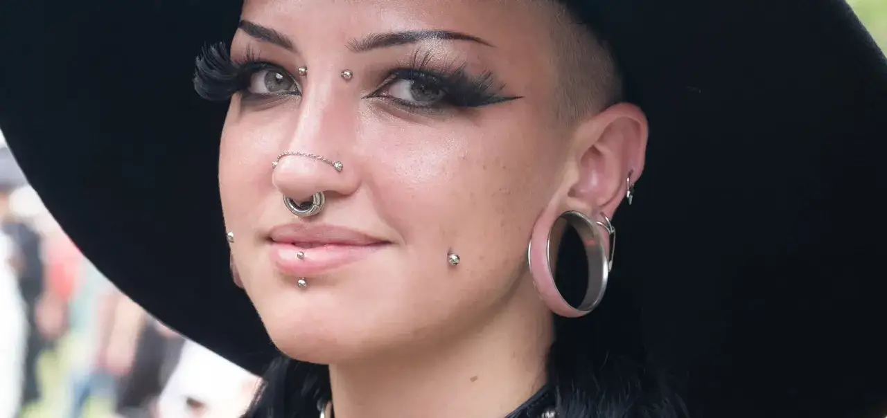 Kobieta z licznymi kolczykami, w tym bridge piercing, septum i kolczykami w policzkach, nosi duży czarny kapelusz.