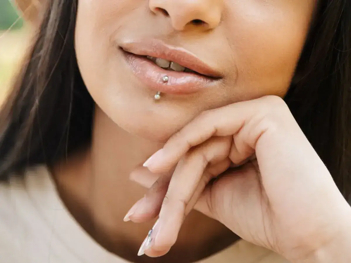 Uśmiechnięta twarz z błyszczącymi ustami i subtelnym ashley piercingiem. Dłoń podparta pod brodą.