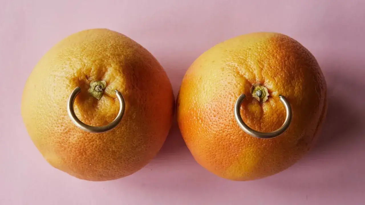 Dwa grejpfruty z metalowymi kółkami, przypominającymi nipple piercing, na różowym tle.