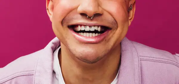 Uśmiechnięta twarz z kolczykiem w nosie i zębami ozdobionymi biżuterią. Widoczny frenum piercing dodaje charakteru.