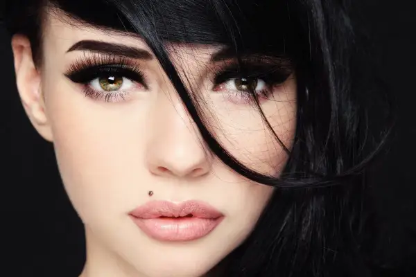Kobieta z czarnymi włosami i makijażem podkreślającym oczy. Widoczny monroe piercing nad górną wargą.